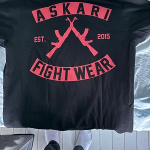 Svart t-shirt Askari Fight Wear M - Svart t-shirt från Askari Fight Wear med stort rött tryck på ryggen och mindre tryck på bröstet. Motivet har två korsade vapen och texten 'ASKARI FIGHT WEAR EST. 2015'. Tillverkad i 100% bomull, rund hals och korta ärmar. Perfekt för dig som gillar kampsport och streetwear.