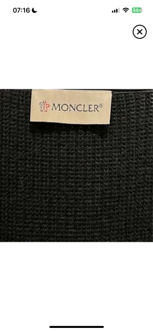 Svart Moncler pufferjacka med huva - Svart pufferjacka från Moncler med stickade ärmar och quiltad front. Jackan har en stor, vadderad huva och dragkedja framtill med Moncler-logga. Snygg kombination av syntet och stickat material, perfekt för höst och vinter. Fickor med dragkedja på ärmen och sidan.