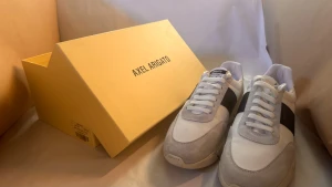 Axel Arigato vita sneakers med grå detaljer - Snygga sneakers från Axel Arigato i vitt tyg med grå mockadetaljer och en svart sidopanel. Klassisk siluett med rund tå, platt sula och snörning. Perfekta för dig som gillar stilrena och trendiga skor med premiumkänsla.  Skorna är endast använda en gång och är i nyskick. Tack vare de neutrala färgerna passar de till alla stilar och kan enkelt matchas med många olika outfits och färger – ett riktigt mångsidigt val oavsett stil. Original boxen följs med om så önskas. Priset är inte hugget i sten!