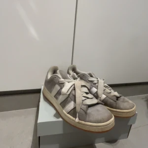 Grå Adidas Campus sneakers mocka - Säljer ett par grå Adidas Campus sneakers i mocka med vita ränder och vit sula. Klassisk låg modell med rund tå och snörning. Snygg retrostil och passar perfekt till streetwear eller casual outfits.