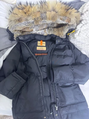 Svart Parajumpers dunjacka med päls - Säljer en svart Parajumpers dunjacka med fluffig päls på huvan och cool gul detaljrem. Jackan har patch på ärmen, dragkedja och stora fickor. Perfekt för kalla vinterdagar och riktigt snygg streetstil. Jackan har en skada vid luvan (se sista bilden). Utöver det finns inga skador. Köptes för 10 500kr hos jackie 