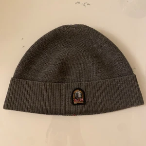 Grå ribbad mössa från Parajumpers - Snygg grå mössa från Parajumpers med ribbstickad uppvikt kant och klassisk PJS-logga framtill. Mjuk och varm, perfekt för kyliga dagar. Materialet är stickad syntet och designen är enkel men stilren, passar till de flesta jackor och stilar. Bara att skriva vid frågor och priset går såklart att diskutera!