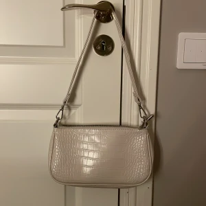 Beige axelväska från Gina Tricot - Snygg beige axelväska från Gina Tricot med croco-mönstrad yta och silvriga metalldetaljer. Väskan har dragkedja upptill och ett rymligt fack med innerficka. Perfekt stor. Nyskick. Endast använd ett fåtal gånger. Några år gammal i årsmodellen. Köparen står för frakten!