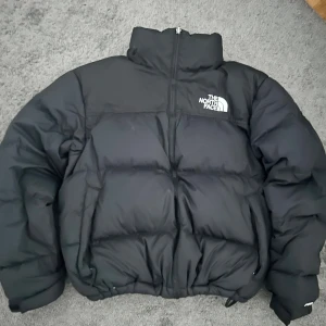 Svart pufferjacka från The North Face - Säljer en svart pufferjacka från The North Face med hög krage och tydlig logga på bröstet och ryggen. Jackan har dragkedja framtill, snörning i nederkant och är rejält vadderad för extra värme. Perfekt för kalla dagar och har en clean, urban look.