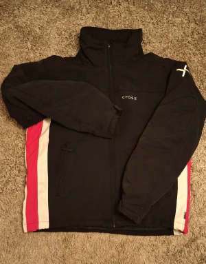 Svart vindjacka från Cross - Svart vindjacka från Cross med dragkedja framtill och högt krage. Jackan har vita och röda paneler längs sidorna och en vit Cross-logga på ena ärmen. Två fickor med dragkedja och sportig look, perfekt för utomhusaktiviteter.