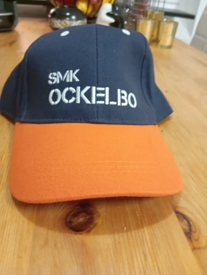 SMK Ockelbo keps marinblå och orange - Snygg keps med marinblå krona och orange skärm. Framtill är kepsen broderad med texten 'SMK OCKELBO' i vitt. Klassisk form med böjd skärm, perfekt för dig som vill sticka ut med färg. Passar till både chill och aktivitet.