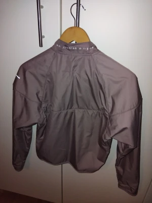 Gammaldagsrosa vindjacka Nike Running Division - Endast provad. Säljer en gammaldagsrosa vindjacka från Nike Running Division med hel dragkedja framtill och hög krage. Jackan har långärmad design och är tillverkad i lätt syntetmaterial, perfekt för löpning och utomhusaktiviteter. Snygg och sportig look med diskreta reflexdetaljer.