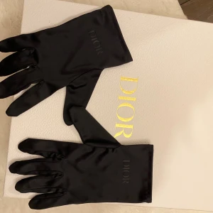 Svarta sidenhandskar från Dior - Exklusiva svarta handskar från Dior i mjukt och glansigt sidenmaterial. Diskret Dior-logga ton-i-ton på ovansidan. Handskarna har en elegant och smal passform som ger en lyxig känsla. Perfekta för dig som vill ha en stilren accessoar med designer-touch. 