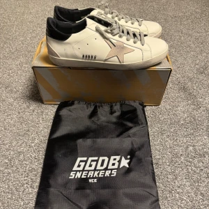 Golden Goose Superstar sneakers vit/svart - Golden Goose Superstar sneakers i vitt skinn med svart häl och grå mockadetaljer. Klassisk stjärna på sidan i ljus mocka, grå snörning och platt sula. Snygg och stilren modell som passar till allt. Levereras med originalkartong och dustbag. Pris kan diskuteras!!!