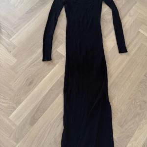 Svart ribbad långklänning från Amisu - Svart långklänning från Amisu i ribbad stickad kvalitet. Klänningen har lång ärm, fyrkantig urringning och en snygg slits framtill. Perfekt för dig som gillar minimalistisk och stilren look med lite edge.