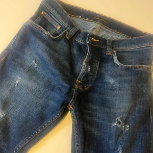 Grim trim jeans nudie  - Grim trim Snygga blå jeans från Nudie Jeans Co med slitna detaljer och orange kontrastsömmar. Klassisk femficksmodell med vågig brodyr på bakfickorna och läderpatch i midjan. Jeansen har en rak passform och är tillverkade i mjukt denimtyg.