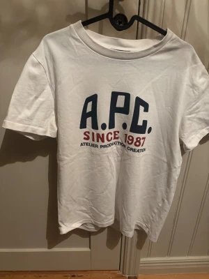 Vit A.P.C. t-shirt med tryck - Säljer en vit t-shirt från A.P.C. med stort tryck på bröstet i blått och rött. Modellen är klassisk med rund hals och korta ärmar. Tillverkad i mjuk bomull och har en clean vibe som passar perfekt till jeans eller shorts.