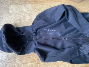 Svart hoodie från Sail Racing med huva - Svart hoodie från Sail Racing med dragkedja hela vägen och en extra zipficka på bröstet. Mjuk fleece på insidan och justerbar huva. Snyggt tryck på bröstet och ribbade muddar vid ärmslut och nederkant. Perfekt för chill eller när du vill vara extra bekväm.