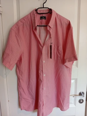 Röd mönstrad skjorta Carl William XXL - Snygg röd skjorta från Carl William i storlek XXL med diskret diamantmönster. Skjortan har korta ärmar, klassisk krage och vita knappar med röda detaljer. Bröstficka på vänster sida. Perfekt för dig som gillar stilrena detaljer och mönster.