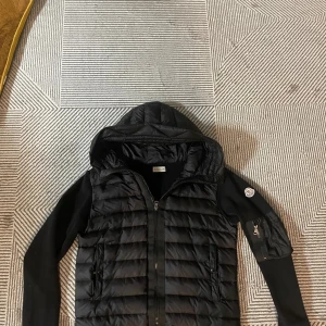 Svart Moncler pufferjacka/cardigan med huva - Svart pufferjacka från Moncler med huva och ribbstickade ärmar. Jackan har dragkedja framtill, två sidofickor med dragkedja och en extra ficka med dragkedja på ärmen. Klassisk Moncler-logga på vänster ärm. Perfekt för kalla dagar och snygg streetstil.