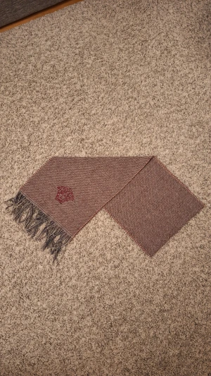 Vinröd halsduk från Versace med fransar - Säljer denna snygga halsduk från Versace med rutig mönster i beige, brun och vinröd. 100% ull. Använd fåtal gånger och skicket är i princip som ny, inga slitningar eller defekter finns. Den har Versace Medusa-loggan broderad i vinrött på båda sidorna. Den är stilig och varm och passar perfekt för dig som vill ha en lyxig touch på outfiten nu under vintern och Jul. Skick: 9/10