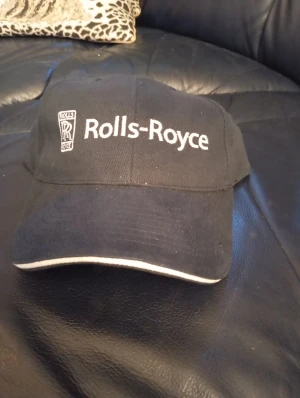 Svart Rolls-Royce keps - Snygg svart keps med vit Rolls-Royce-logga broderad framtill. Klassisk böjd skärm med vit kant och justerbar passform. Perfekt för dig som gillar exklusiva bilar och vill ha en stilren accessoar. Materialet känns som bomull och kepsen har en tidlös look.