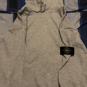 Grå hoodie från Stone Island - Säljer en stilren grå hoodie från Stone Island med klassisk patch på vänster ärm. Tröjan har huva med snörning och är tillverkad i mjuk bomull. Perfekt för dig som gillar streetwear och vill ha något bekvämt och snyggt. Storlek L, priset kan diskuteras.