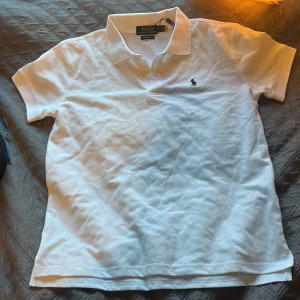 Vit pikétröja Ralph Lauren slim fit - Vit pikétröja från Ralph Lauren i custom slim fit med klassisk krage och två knappar. Tröjan har den ikoniska broderade loggan på bröstet och är gjord i 100% bomull. Perfekt för dig som gillar stilrena och tidlösa plagg.