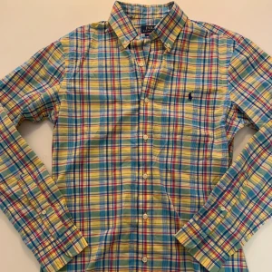 Rutig skjorta Polo Ralph Lauren S - Färgglad rutig skjorta från Polo Ralph Lauren i slim fit. Skjortan har långa ärmar, klassisk krage och knappar framtill. Materialet är mjuk bomull och mönstret går i blått, gult, rött och grönt. Ikonisk broderad logga på bröstet.