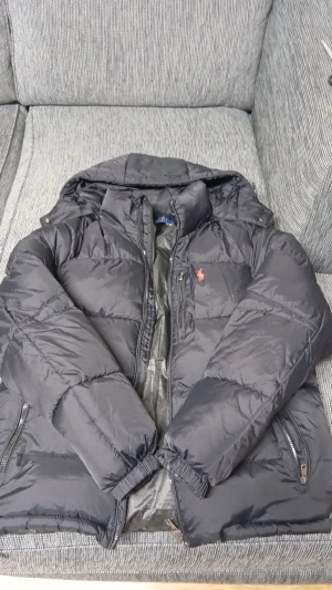 Svart pufferjacka Polo Ralph Lauren M - Svart pufferjacka från Polo Ralph Lauren i storlek M. Jackan har broderad röd logga på bröstet, dragkedja framtill och två sidofickor med dragkedja. Klassisk pufferstil med hög krage och vadderad känsla. Perfekt för kalla dagar.