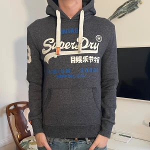 Mörkgrå Superdry hoodie - Riktigt snygg superdry hoodie. Modellen är 187 och hoodien är i storlek S. Hör av er vid minsta funderingar🤗