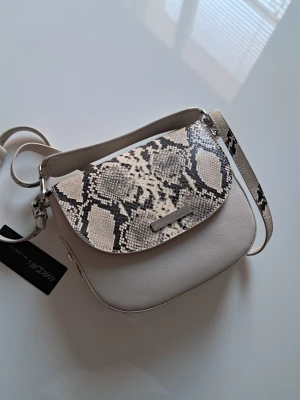 Axelväska i skinn från Marc Cain - Snygg axelväska från Marc Cain i ljusbeige skinn med coolt ormmönstrat lock och axelrem. Väskan har en rundad form och silverfärgad logodetalj framtill. Insidan är fodrad med polyester. Perfekt för dig som vill sticka ut med en trendig accessoar. (23×19×5)cm