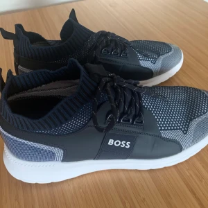 Run Extreme Knit från BOSS - Snygga sneakers från BOSS med stickad ovandel i svart och vitt, vita platta sulor och snörning. Skorna har en modern, sportig design med rund tå och BOSS-logga på sidan. Materialet är en mix av syntet och stickad textil för skön komfort.