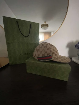 Gucci beige keps med GG-mönster - Snygg beige keps från Gucci med klassiskt GG-monogram över hela kepsen och en ikonisk grön-röd rand på sidan. Tillverkad i bomull och polyester, med brunt innerfoder och justerbar rem baktill. Perfekt för dig som vill ha en lyxig streetstyle-accessoar.