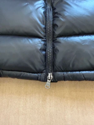 Svart pufferjacka från Polo Ralph Lauren - Svart pufferjacka från Polo Ralph Lauren med huva, dragkedja och två sidofickor. Jackan har en röd Polo-logga på bröstet och justerbara snören vid nederkant. Materialet är glansigt och syntetiskt, perfekt för kalla dagar.