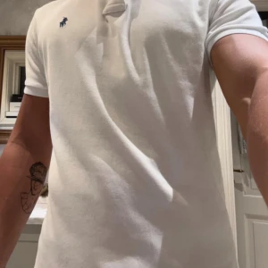 Vit pikétröja från Polo Ralph Lauren - Vit pikétröja från Polo Ralph Lauren i slim fit med klassisk krage och två knappar. Tröjan har det ikoniska blå broderade Polo-logot på bröstet och är gjord i mjuk bomull. Perfekt för dig som gillar stilrena och tidlösa plagg.