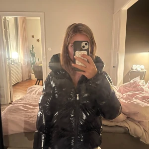 Svart glansig pufferjacka från Colmar - Säljer en svart, glansig pufferjacka från Colmar med huva och dragkedja framtill. Jackan har en quiltad look och är perfekt för kalla dagar. Klassisk passform och diskret logga på ärmen. Riktigt snygg och trendig vinterjacka!