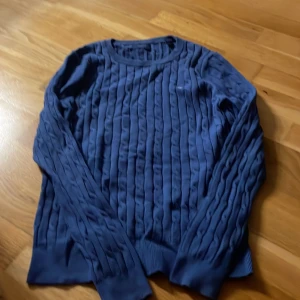 Marinblå kabelstickad tröja från GANT - Snygg marinblå kabelstickad tröja från GANT med rund hals och diskret logga på bröstet. Tröjan har långärmad passform och ribbade muddar vid ärmslut och nederkant. Perfekt för dig som gillar klassisk stil med en modern twist.