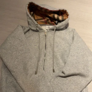 Grå hoodie från Burberry med dragkedja - Snygg grå hoodie från Burberry i storlek M med klassiskt rutigt foder i huvan. Tröjan har dragkedja framtill, snörning vid huvan och subtilt Burberry-broderi på bröstet. Perfekt för dig som gillar stilrena och lyxiga detaljer. Priset kan diskuteras!