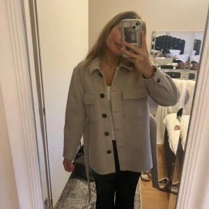 Beige kappa med bälte och stora fickor - Säljer en beige kappa med stora svarta knappar och två rejäla bröstfickor. Jackan har ett bälte i midjan som kan knytas för en snygg siluett. Materialet känns mjukt och lite ulligt. . Långärmad och med klassisk krage. Passar xs och s