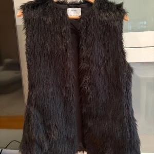 Svart pälsväst från Bershka - Snygg svart pälsväst från Bertha med lång, fluffig fuskpäls och rak passform. Västen är ärmlös och har en öppen front utan knäppning, vilket ger en cool och avslappnad look. Perfekt att slänga över en hoodie eller tröja för extra edge.