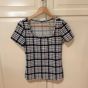 Rutig topp med fyrkantig hals - Snygg topp med rutigt mönster i svart, vit och blå. Modellen har en fyrkantig halsringning och korta ärmar. Materialet känns mjukt och stretchigt.