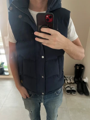 Mörkblå dunväst Superdry Everest - Snygg mörkblå dunväst från Superdry Everest med huva och stora fickor. Västen har både dragkedja och tryckknappar framtill, samt en diskret Superdry-logga vid fickan. Perfekt för lager-på-lager och chill stil under kyliga dagar. Nypris 1399 mitt pris 999kr. Aldrig använd. 