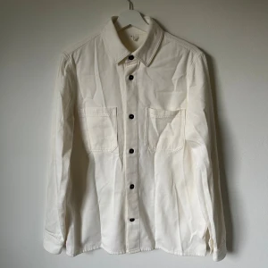 Vit Arket overshirt i ekologisk denim - Overshirt från Arket i mjuk, ekologisk bomullsdenim. Skjortan är vit med svarta knappar, har två stora bröstfickor och klassisk krage. Långärmad modell med knappslå vid ärmslut. Perfekt för lager-på-lager och clean stil. 5J7