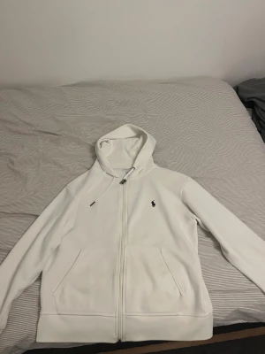 Vit hoodie från Polo Ralph Lauren - Vit hoodie från Polo Ralph Lauren med dragkedja och klassisk logga broderad på bröstet. Tröjan har huva med snörning, två fickor fram och är långärmad. Perfekt för chill dagar och enkel att matcha med jeans eller joggers.