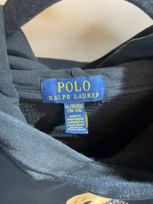 Svart Polo Bear hoodie från Ralph Lauren - Svart hoodie från Ralph Lauren med Polo Bear-tryck på framsidan där björnen håller en present. Tröjan har huva och ribbade muddar. Materialet är mjuk bomull och polyester, perfekt för dig som gillar ikoniska motiv och chill stil.
