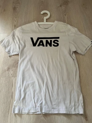 Vit Vans t-shirt med logga - Vit t-shirt från Vans i klassisk passform med stor svart Vans-logga på bröstet. Tillverkad i 100% bomull, rund hals och korta ärmar. Perfekt för dig som gillar streetwear och enkel stil.