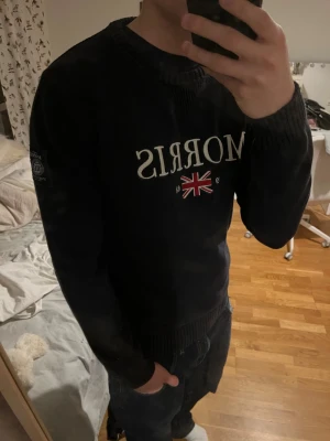 Svart/mörkblå stickad tröja Morris M - Svart/mörkblå stickad tröja från Morris med broderad logga och brittisk flagga framtill. Tröjan har ribbade muddar vid hals, ärmslut och nederkant samt en patch på vänster ärm. Snygg och skön tröja! Inga defekter och perfekt för kallare dagar. Storleken är M och hör av dig gällande frågor!!