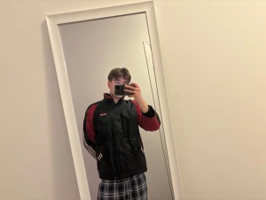 Svart och röd Adidas vindjacka - Adidas vindjacka i svart och rött med klassiska vita ränder på ärmarna och logga på bröstet. Jackan har hög krage, snörning i midjan, dragkedja och knappar framtill. Tillverkad i polyester och har två fickor med dragkedja. Perfekt för dig som gillar sportig stil.