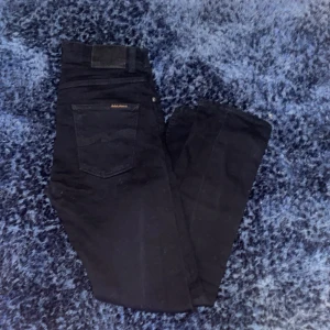 Svarta jeans från Nudie Jeans - Säljer ett par svarta jeans från Nudie Jeans i storlek 32/32. Klassisk femficksmodell med raka ben och normal passform. Jeansen har detaljer som nitar och läderpatch bak i midjan.