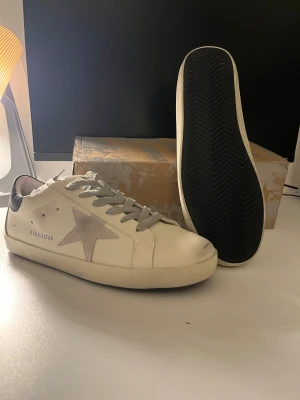 Golden Goose vita sneakers med stjärna  - Golden Goose sneakers i vitt skinn med grå mockadetaljer och ikonisk stjärna på sidan. Grå snörning, svart hälparti och mörkblå mocka baktill. Klassisk låg modell med platt sula och GGDB/SSTAR-tryck på sidan. Perfekta för en cool och avslappnad stil.