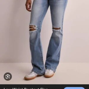 Bootcut ljusblå jeans med slitningar - Säljer ett par ljusblå bootcut jeans med snygga slitningar och hål vid knäna. Jeansen har klassiska fickor och är tillverkade i jeansmaterial med en låg midja. Perfekt för dig som gillar en avslappnad och trendig stil.