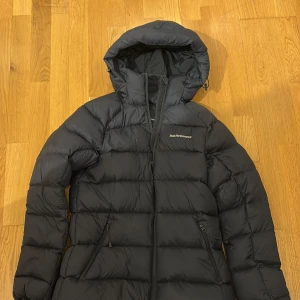 Svart Peak Performance dunjacka - Säljer en svart dunjacka från Peak Performance med huva och dragkedja framtill. Jackan har två praktiska fickor med dragkedja. Perfekt för kalla dagar och har en snygg, normal passform.