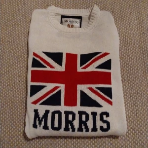 Vit stickad tröja Morris - Vit stickad tröja från Morris med stor flagga i rött, blått och vitt på bröstet samt Morris-logga under. Tröjan har långärmad passform och två mörkblå ränder på ena ärmen. Rund hals och ribbade muddar ger en klassisk look. Riktigt skön tröja som passar mig som är 180 74kg, inga defekter och storleken är L men passar mig som är M så skulle säga att den är M. Hör av dig gällande frågor😎😎