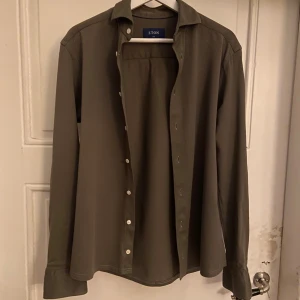 Olivgrön skjorta från Eton  - Riktigt schysst olivgrön skjorta från Eton!Skjortan har klassisk krage, vita knappar och långa ärmar. Materialet är mjukt med 100% luftigt bomull, vilket gör den skön att bära hela dagen. 